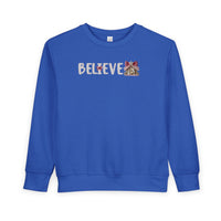 Royal-blue toddler crewneck sweatshirt with BELIEVE in white knit-look font, featuring a nativity scene as the final E and a red bow on the I.