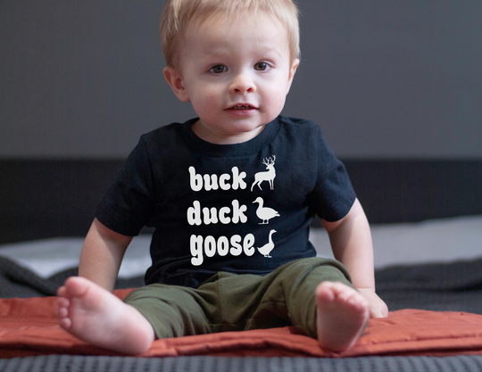 Toddler wearing the Buck, Duck, Goose Baby Tee, featuring playful animal silhouettes and text, paired with olive pants. The shirt is soft, shrink-resistant, and durable.