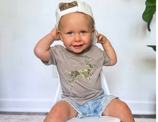 Toddler wearing a Camo Deer Toddler Tee with a camouflage deer print, paired with denim shorts and a white cap, seated on a white chair.