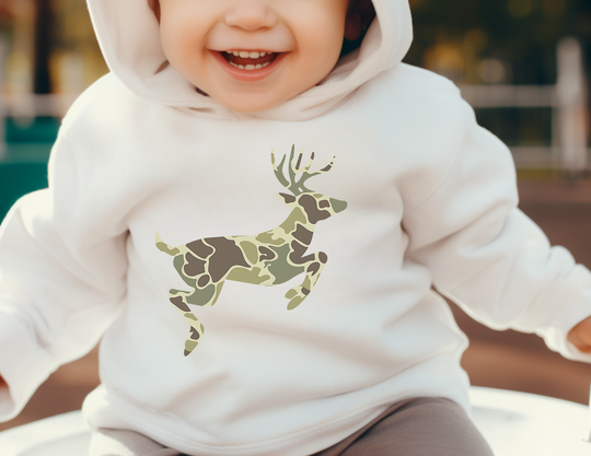 Toddler smiling in a Camo Deer Hoodie, featuring a camouflage deer silhouette. The hoodie offers comfort with its jersey-lined hood and side-seam pockets for coziness.