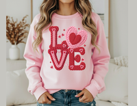 Person wearing Love Heart Crew sweatshirt with LOVE in chunky, braided yarn-style graphic and tiny hearts, paired with blue jeans.