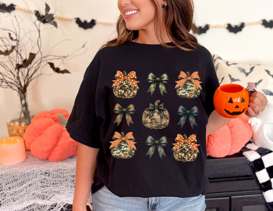 Person wearing the Bows and Pumpkins Tee, featuring a 3x3 grid of stylized pumpkins and bows, holding a jack-o’-lantern mug.
