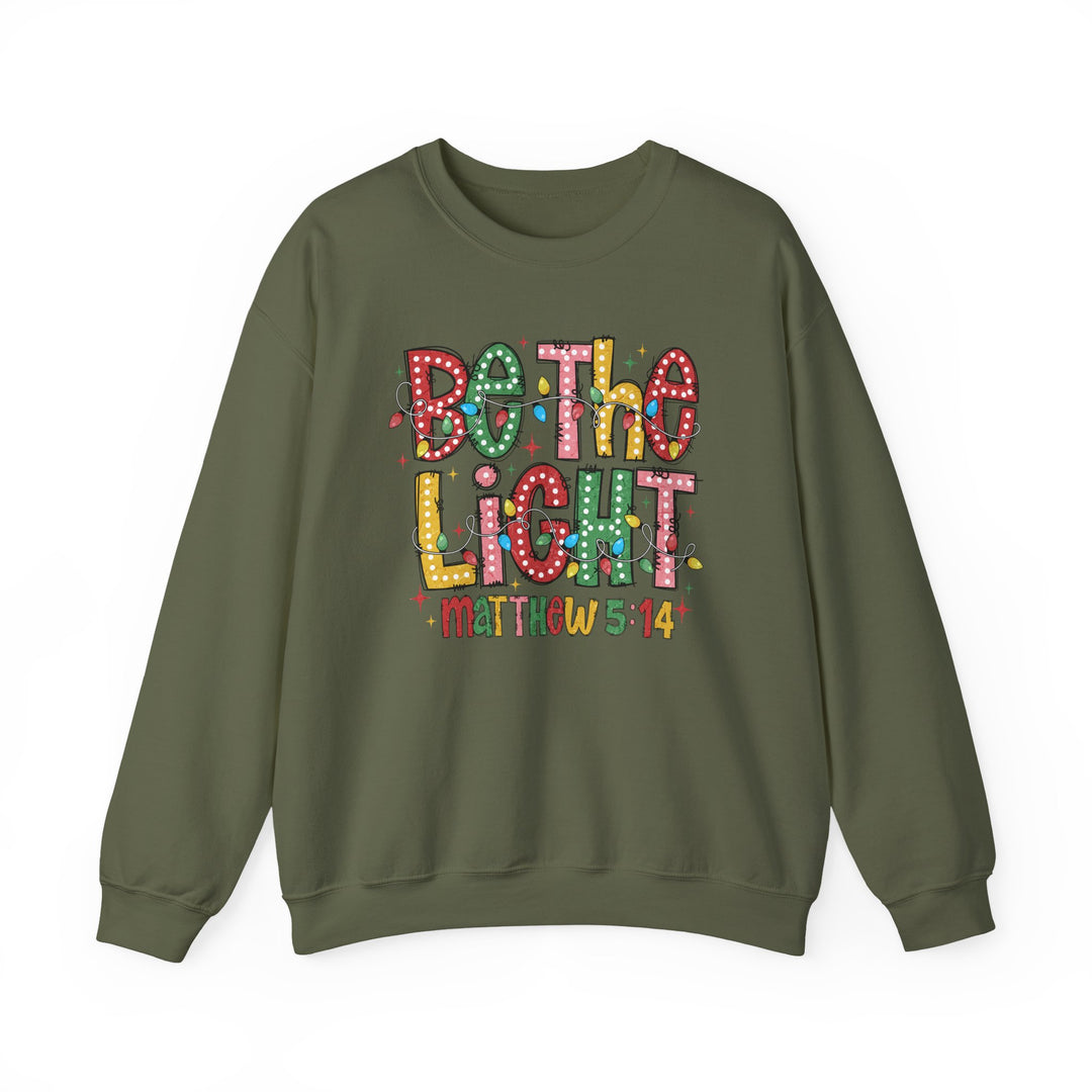 Olive-green sweatshirt with “Be The Light” and Christmas lights design, featuring “Matthew 5:14” below. Cozy, durable, and perfect for colder months.