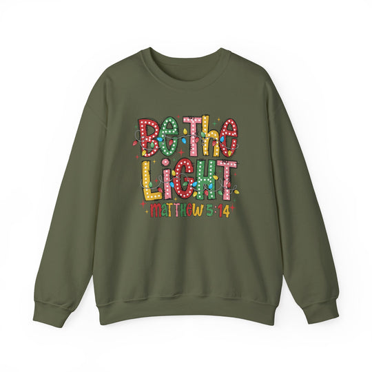 Olive-green sweatshirt with “Be The Light” and Christmas lights design, featuring “Matthew 5:14” below. Cozy, durable, and perfect for colder months.