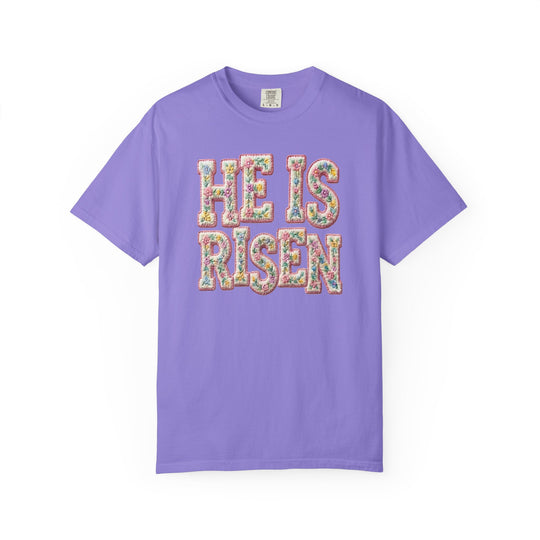 He is Risen Tee in lavender with bold floral-patterned letters and 'Comfort Colors' tag, offering a relaxed fit and soft cotton blend.