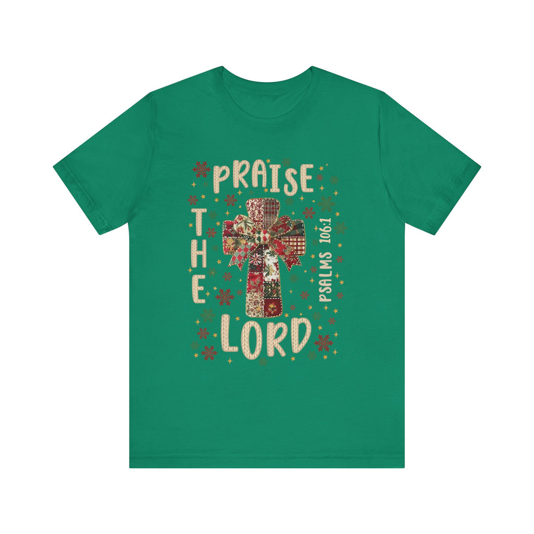 Green T-shirt featuring a patchwork cross with bow, snowflakes, stars, and PRAISE THE LORD text. Bible reference “Psalms 106:1.” Classic unisex fit, soft cotton.