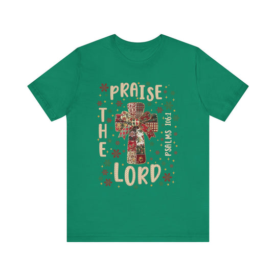 Green T-shirt featuring a patchwork cross with bow, snowflakes, stars, and PRAISE THE LORD text. Bible reference “Psalms 106:1.” Classic unisex fit, soft cotton.