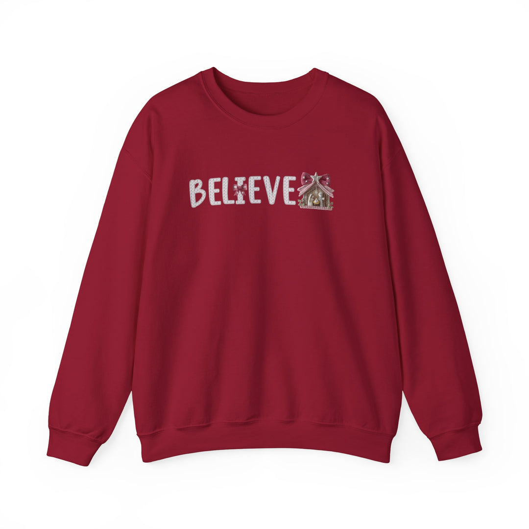 Red crew-neck sweatshirt featuring BELIEVE in candy-cane lettering with a cross-shaped candy I and gingerbread-style nativity stable, ideal for cozy wear.