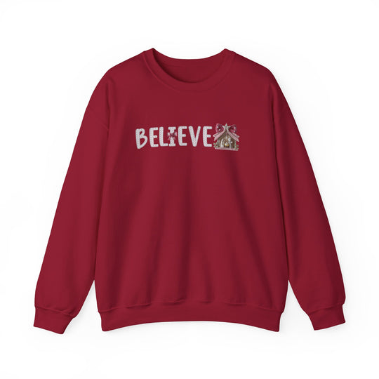 Red crew-neck sweatshirt featuring BELIEVE in candy-cane lettering with a cross-shaped candy I and gingerbread-style nativity stable, ideal for cozy wear.