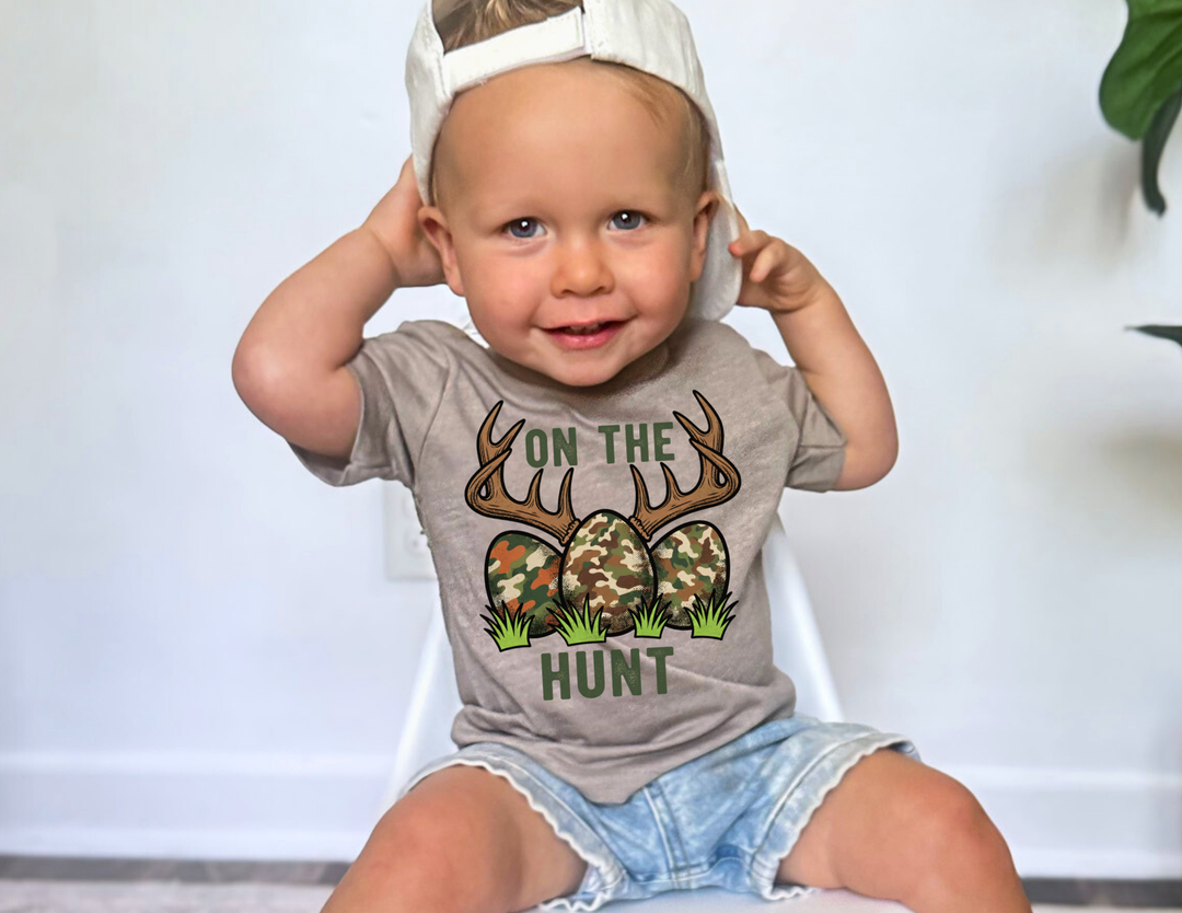 Toddler wearing On the Hunt tee with camouflage eggs and antlers graphic, sitting on chair, smiling, in light-wash denim shorts and backwards cap.