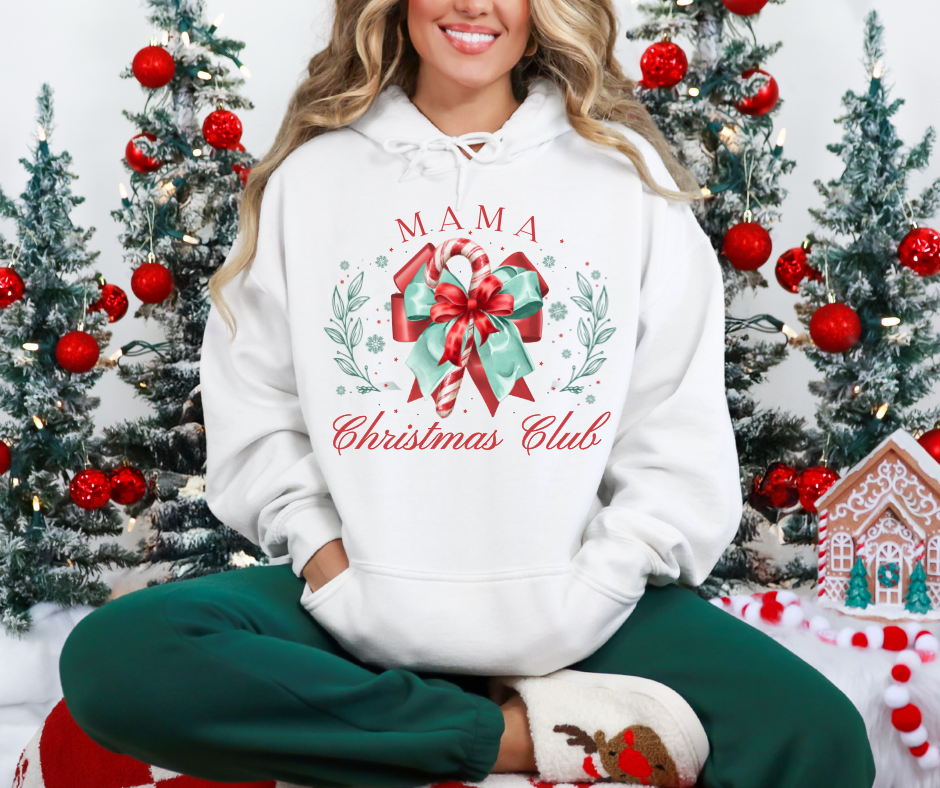 Person wearing MAMA Christmas Club hoodie with candy-cane graphic, dark green lounge pants, and reindeer slippers, seated in a festive setting with decorated Christmas trees.