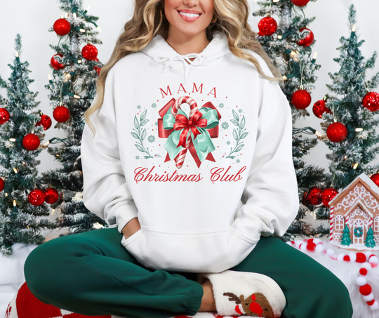 Person wearing MAMA Christmas Club hoodie with candy-cane graphic, dark green lounge pants, and reindeer slippers, seated in a festive setting with decorated Christmas trees.