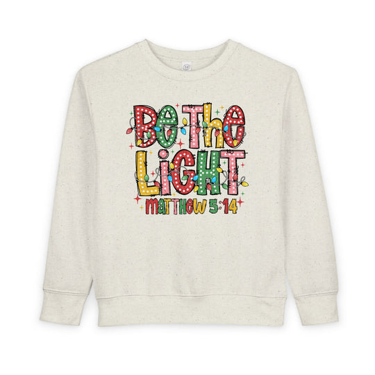 Light-beige toddler sweatshirt with colorful Be the Light graphic resembling Christmas lights, featuring Matthew 5:14 underneath, crafted for warmth and comfort.
