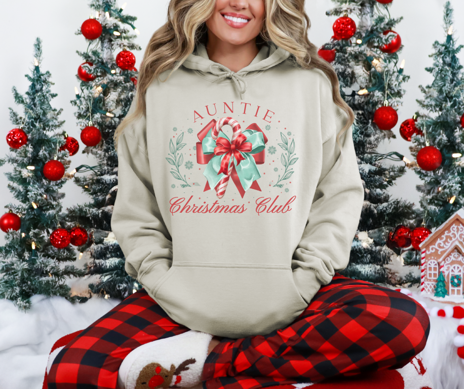 Person in Auntie Christmas Club hoodie, buffalo-plaid pants, and reindeer slippers, sitting amid festive decor with small Christmas trees and a gingerbread house.