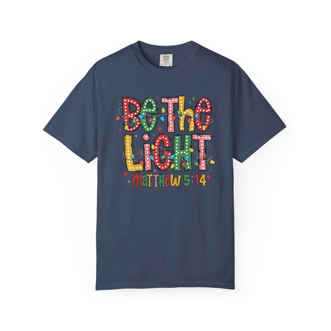 Be the Light Tee featuring Bethlehem Light graphic with festive string lights and Matthew 5:14 text, offering a holiday-themed style.
