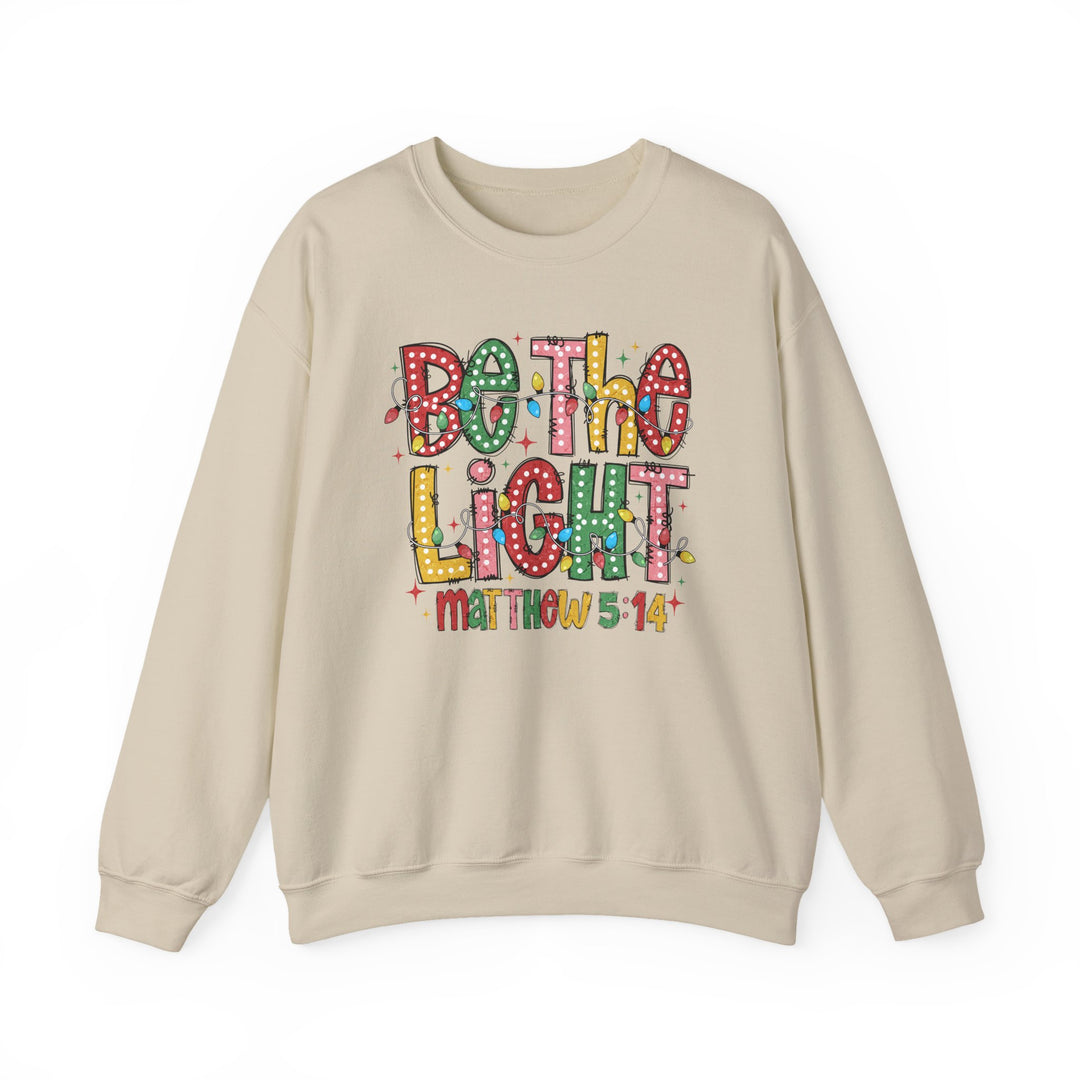 Light-beige crewneck sweatshirt with Be the Light in colorful letters wrapped in string lights, featuring Matthew 5:14 below. Cozy, durable, unisex design.