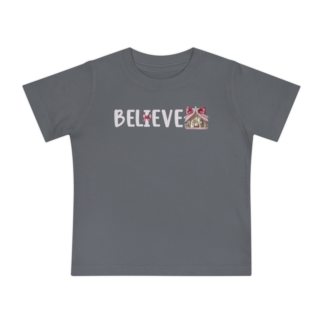 Believe Baby Tee: Slate gray children's T-shirt with BELIEVE in white, featuring a pink-bowed I and gingerbread-style Christmas stable, perfect for stylish first steps.