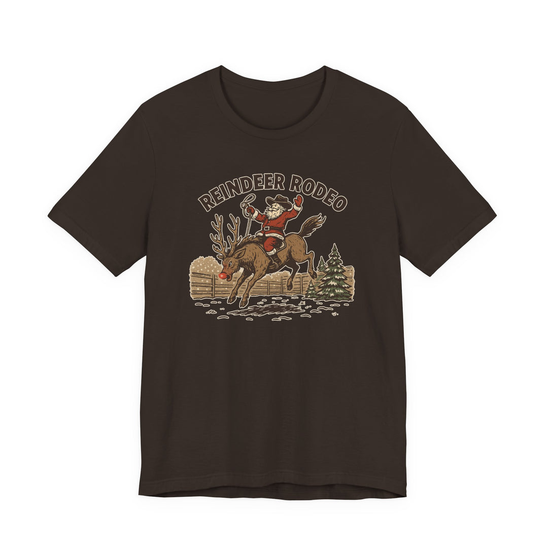 Reindeer Rodeo Tee featuring Santa as a cowboy on a reindeer, set against a snowy backdrop with a fence and pine tree.