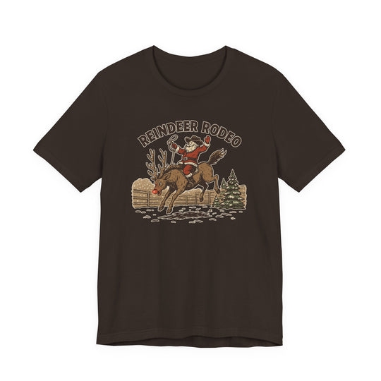 Reindeer Rodeo Tee featuring Santa as a cowboy on a reindeer, set against a snowy backdrop with a fence and pine tree.
