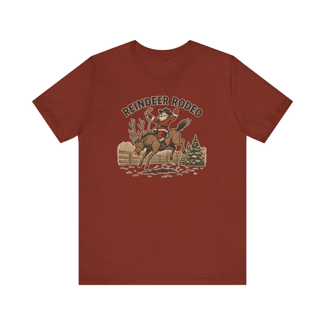 Reindeer Rodeo Tee featuring Santa as a cowboy with lasso, bucking red-nosed reindeer, snowy fence, pine tree, and REINDEER RODEO text, on a rust-colored T-shirt.