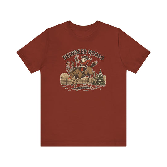 Reindeer Rodeo Tee featuring Santa as a cowboy with lasso, bucking red-nosed reindeer, snowy fence, pine tree, and REINDEER RODEO text, on a rust-colored T-shirt.