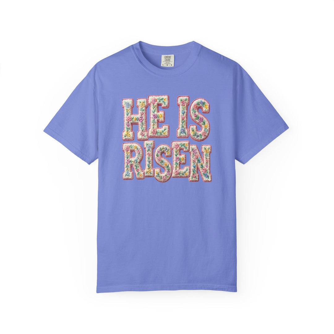He is Risen Tee featuring floral-patterned lettering on lavender-blue, short-sleeved shirt; 'Comfort Colors' label inside collar.