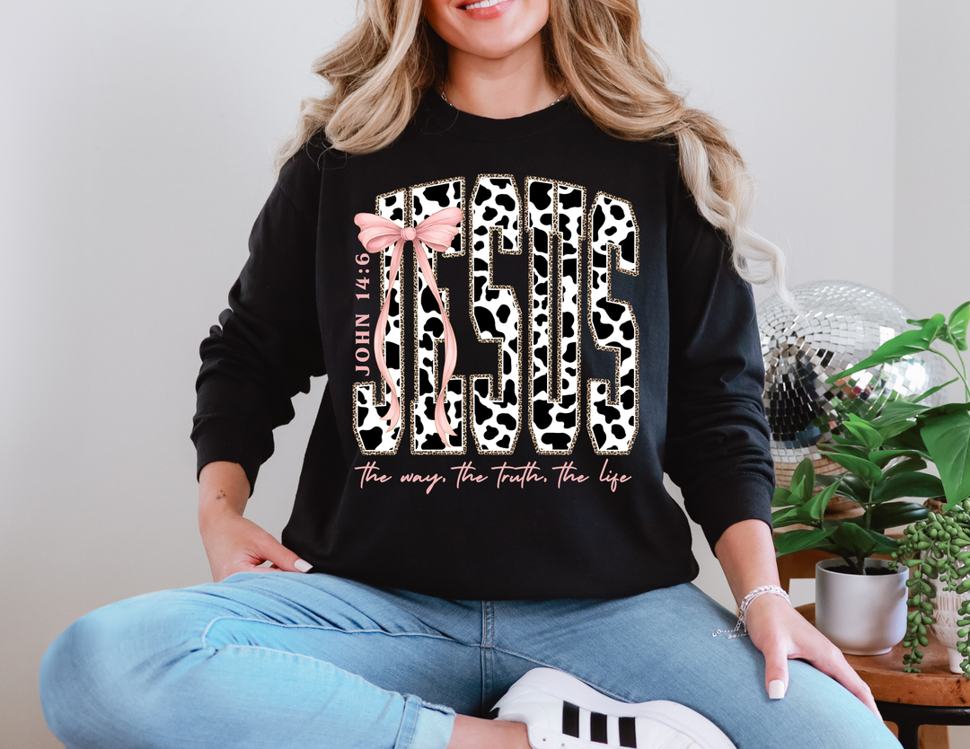 Woman wearing the John 14:6 Long Sleeve Tee with cow-print JESUS design, pink accents, light-wash jeans, and white sneakers, seated beside plants and disco ball.