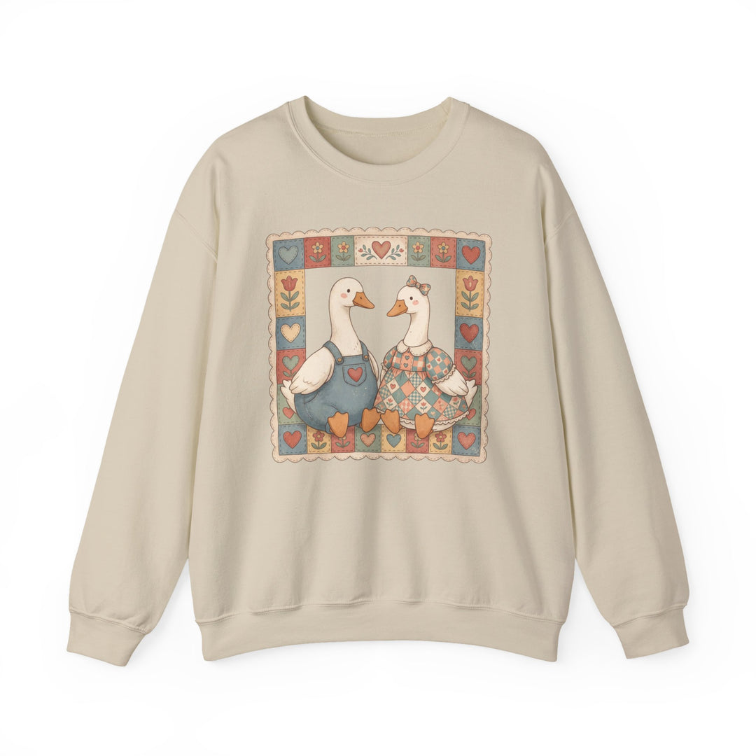 Thrifted Geese Crew: Unisex beige sweatshirt featuring two folk-art geese in clothes, framed by a heart and flower quilt-like border. Cozy, loose fit, cotton-polyester blend.