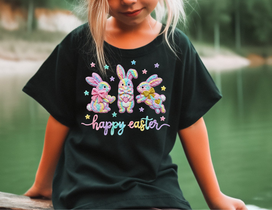 Child wearing Easter Bunny Kids Tee with pastel-rainbow embroidered bunnies and happy easter text, sitting by a lake. Perfect for everyday use.