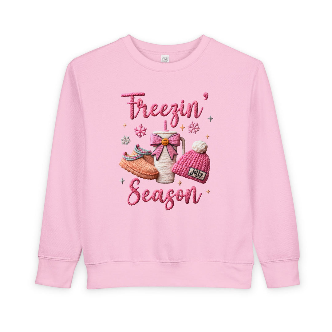 Light-pink Freezin’ Season toddler sweatshirt with embroidered winter-themed design, featuring slippers, a mug, pom-pom hat, snowflakes, and sparkles. Cozy, soft, and durable.