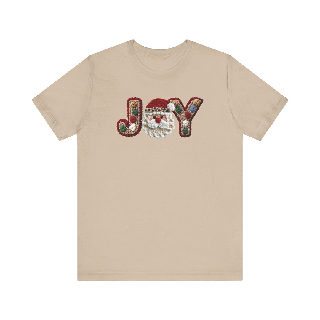 Joy Tee: Light-tan short-sleeve t-shirt featuring JOY with festive hooked-yarn design and Santa Claus face. Soft cotton, unisex fit, durable and comfortable.