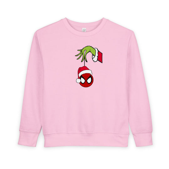 Light-pink toddler sweatshirt features a Grinch-like hand holding a Spider-Man mask ornament. Made from cozy cotton-polyester blend for warmth and comfort.