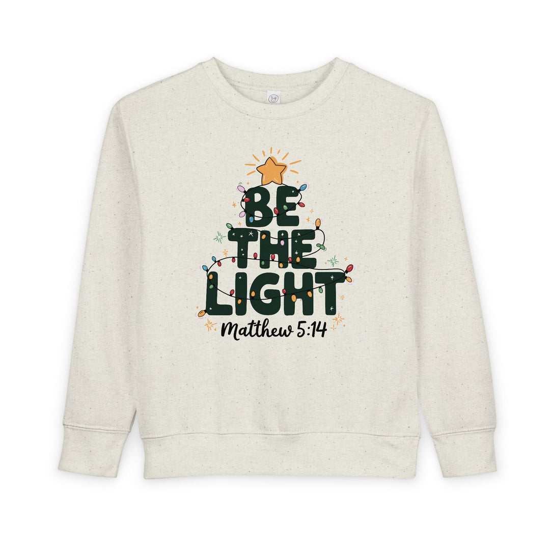 Light-cream toddler crewneck sweatshirt with BE THE LIGHT forming a tree, adorned with colorful lights and a gold star, featuring Matthew 5:14 reference.