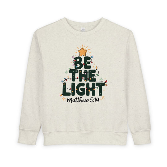 Light-cream toddler crewneck sweatshirt with BE THE LIGHT forming a tree, adorned with colorful lights and a gold star, featuring Matthew 5:14 reference.