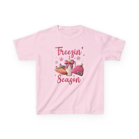 Freezin Season Kids Tee featuring a winter-themed graphic with cozy elements like a mug, slippers, and beanie, on a light-pink, short-sleeve cotton shirt.