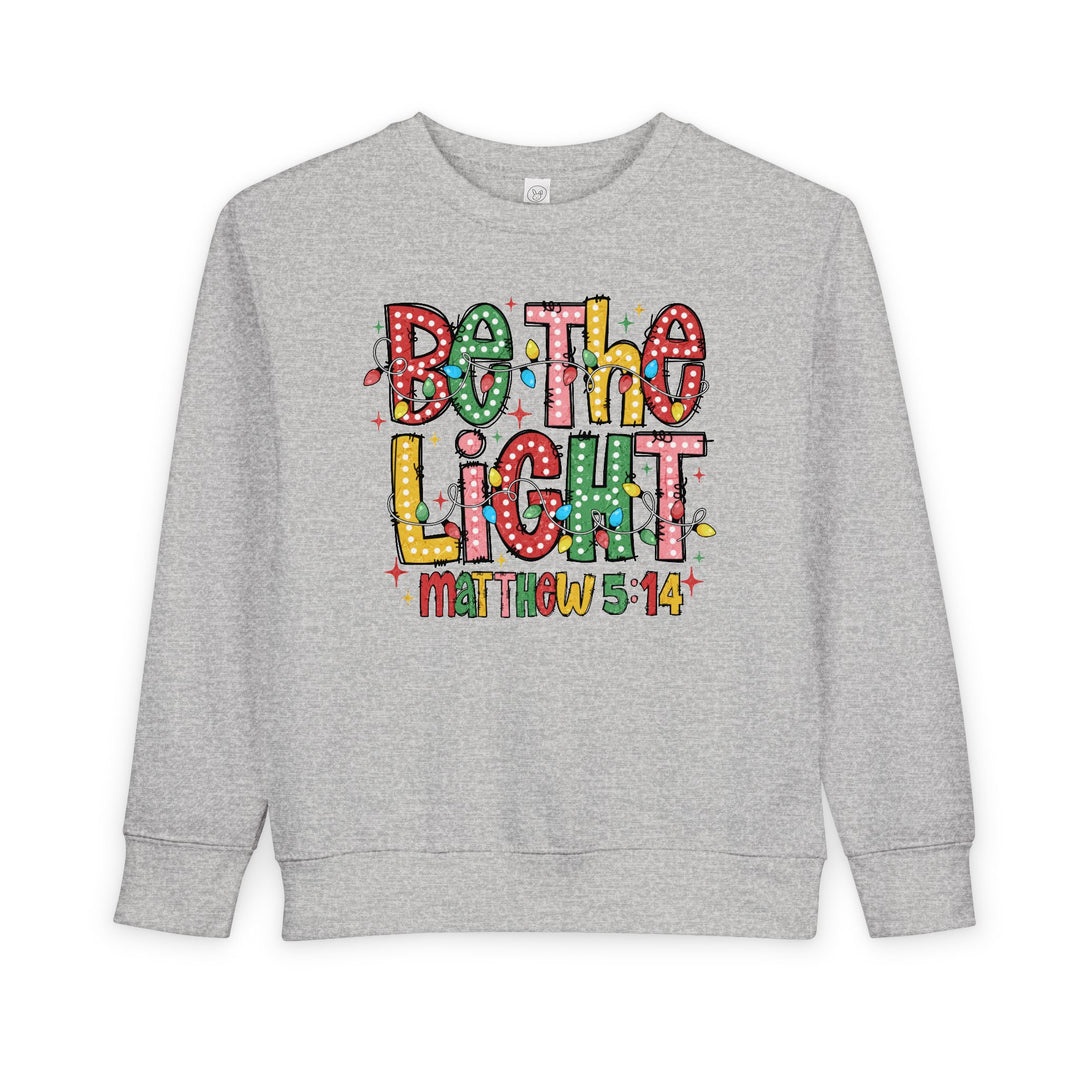 Light-gray toddler crewneck sweatshirt with Be the Light design, featuring colorful, polka-dotted letters wrapped in Christmas lights, highlighting comfort and durability.