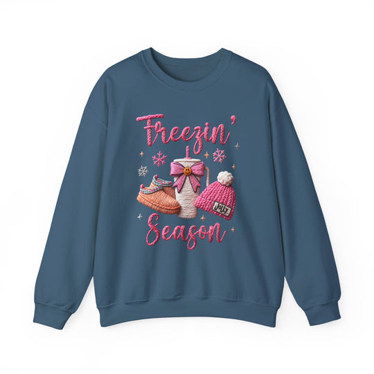 Freezin Season Crew sweatshirt with winter-themed graphic, featuring Freezin’ Season text, slippers, hot drink cup, knit beanie, snowflakes, and sparkles on a blue crewneck.