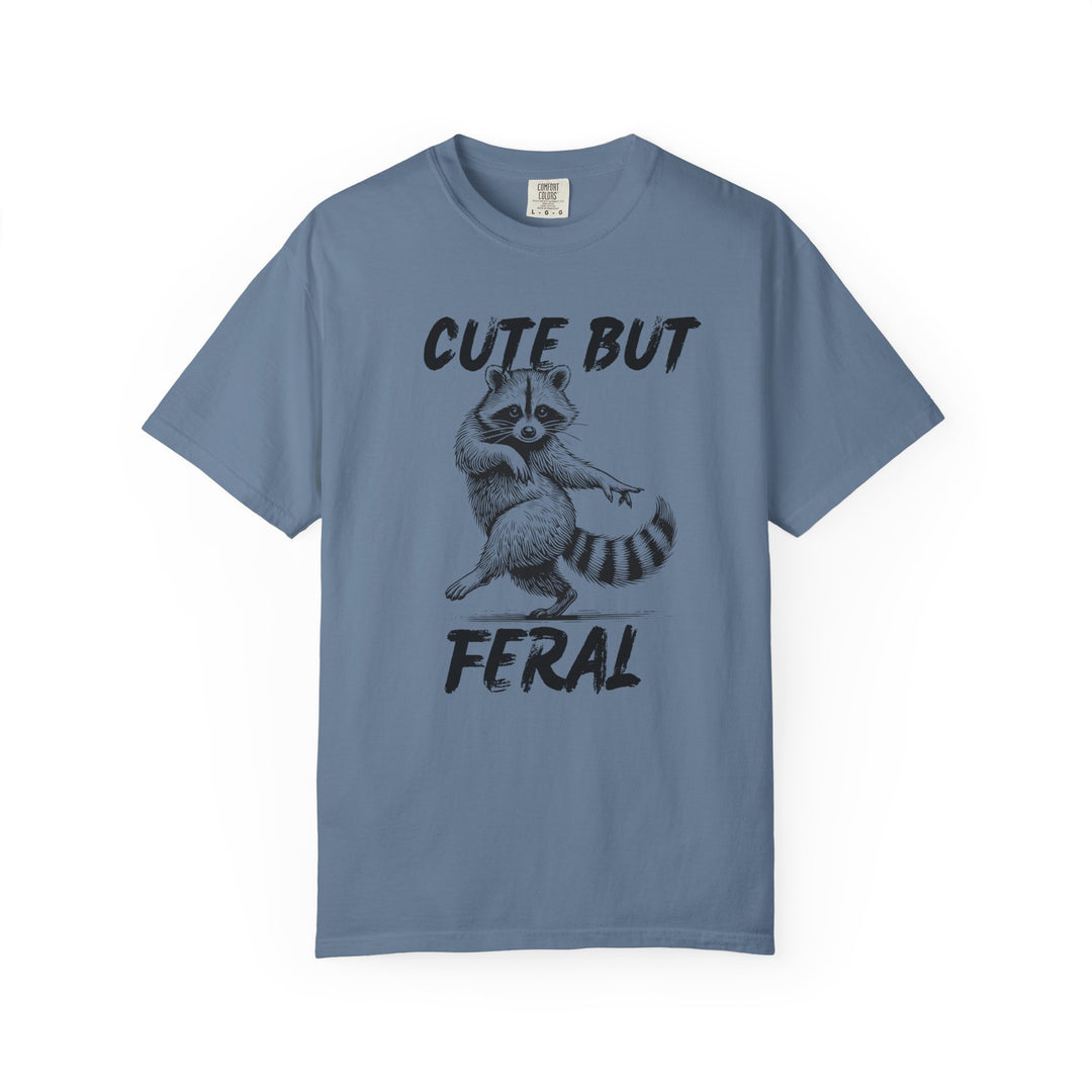 Slate-blue Comfort Colors Cute but Feral tee featuring a black illustration of a dancing raccoon, with bold text above and below, ideal for casual wear.