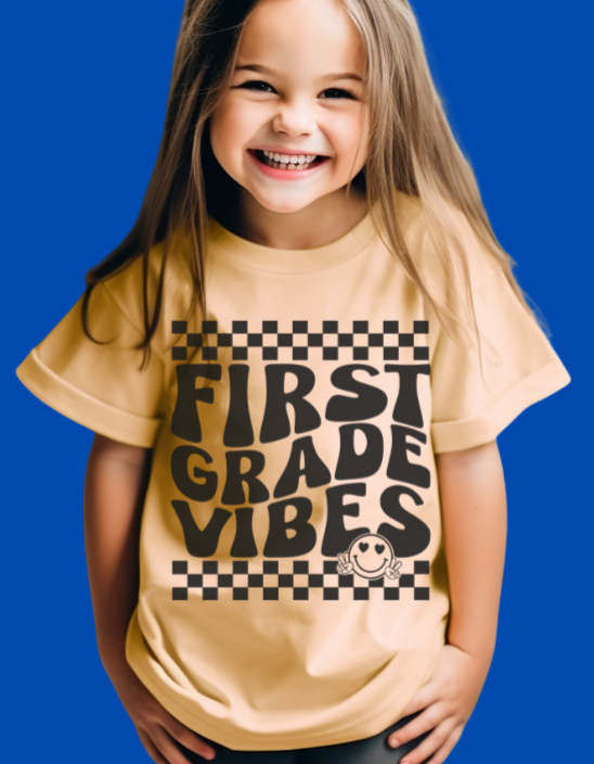 Young girl wearing a FIRST GRADE VIBES yellow T-shirt with checkerboard borders and a peace-sign graphic, embodying playful, durable, everyday wear.