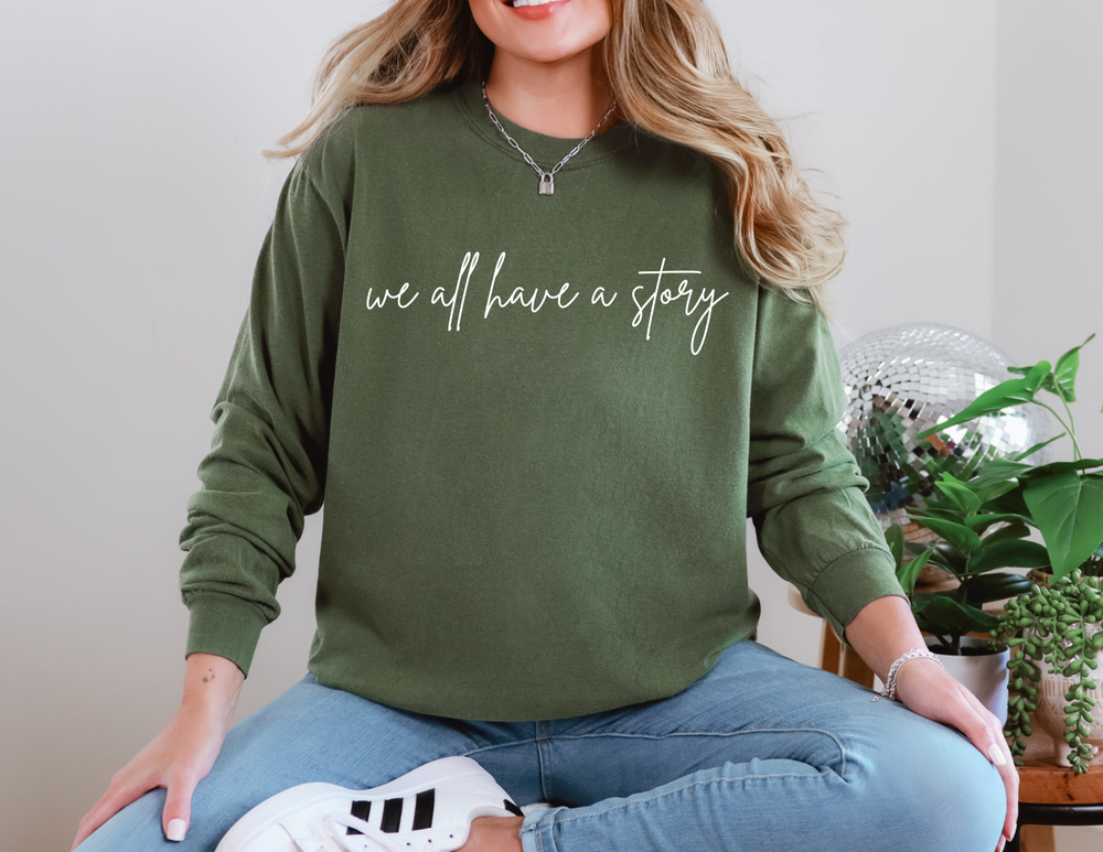 We all have a Story Long Sleeve Tee worn by a woman seated cross-legged, showcasing its relaxed fit and casual style.