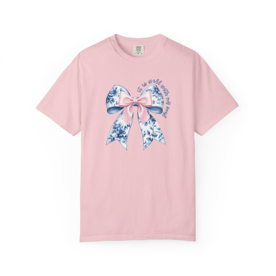 It is Well With My Soul Tee featuring a decorative bow graphic, blending pink and floral patterns, with a comforting phrase in elegant script on a pastel-pink T-shirt.