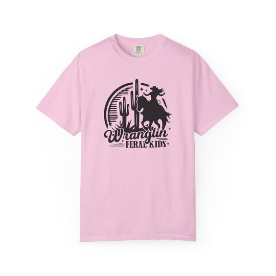 Wranglin’ Feral Kids Tee: Light pink Comfort Colors T-shirt featuring a silhouetted rider on a bucking horse, desert scene, and slogan. Available in sizes S to 4XL.