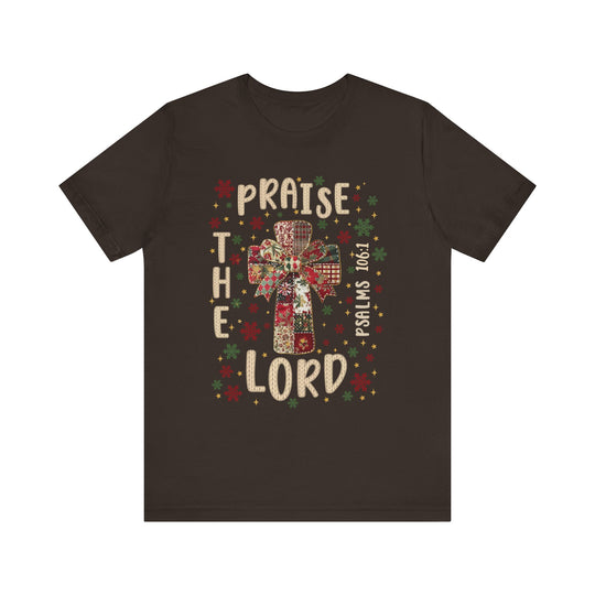 Praise the Lord Tee features a patchwork cross with a bow, surrounded by festive snowflakes and stars, and includes Psalms 106:1 text.