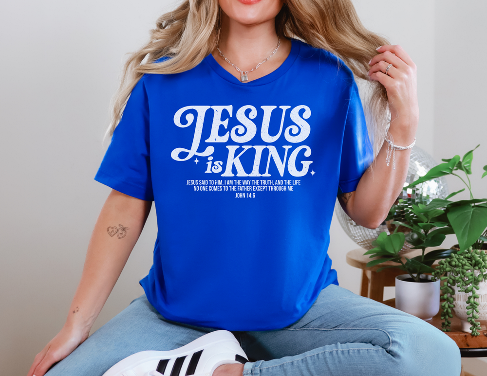 Woman wearing the Jesus is King Tee while seated, highlighting the shirt's casual fit and ribbed knit collar. A plant is visible nearby.