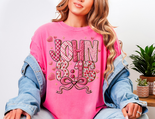Person wearing a 3:16 Tee bright pink sweatshirt over a denim jacket, featuring heart patterns and stars, sitting near a table with potted plants.