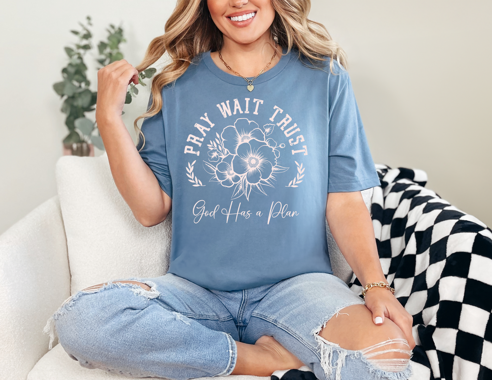 Pray Wait Trust Tee featuring a woman sitting on a couch, wearing casual jeans and a gold bracelet, showcasing the tee's comfortable, stylish fit.