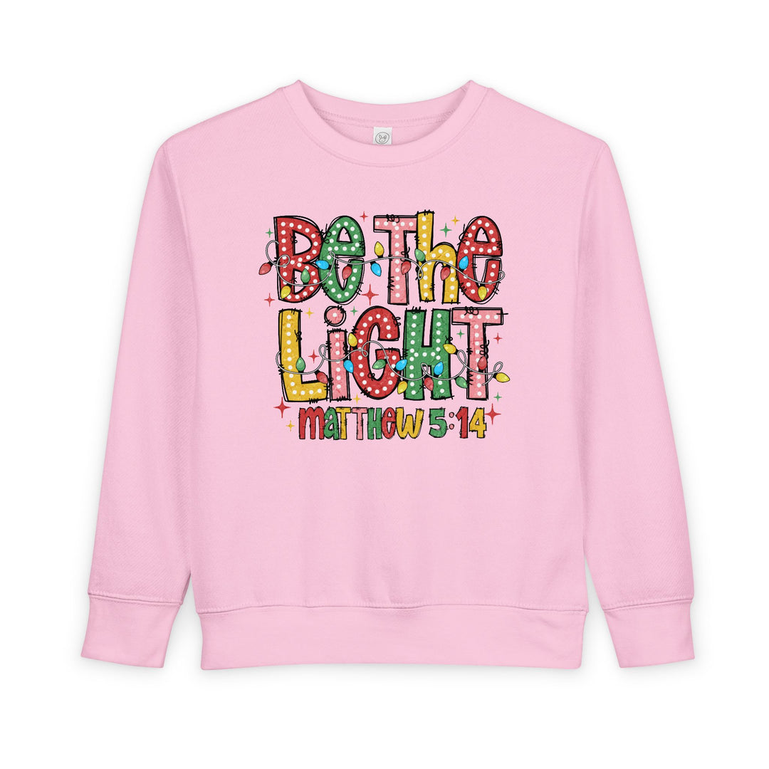 Light-pink toddler crewneck sweatshirt with Be the Light in colorful letters and Christmas lights, featuring Matthew 5:14 reference; cozy cotton-poly fleece blend.