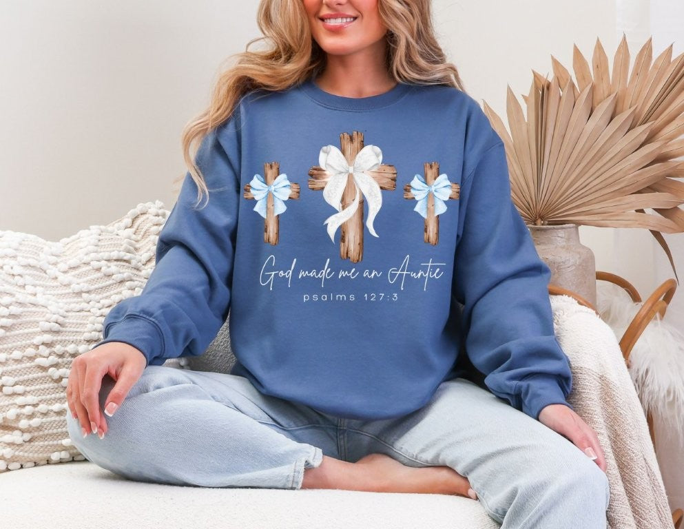 Person in blue God Made Me a Boy Auntie Crew sweatshirt with cross and bow design, seated casually on a couch.