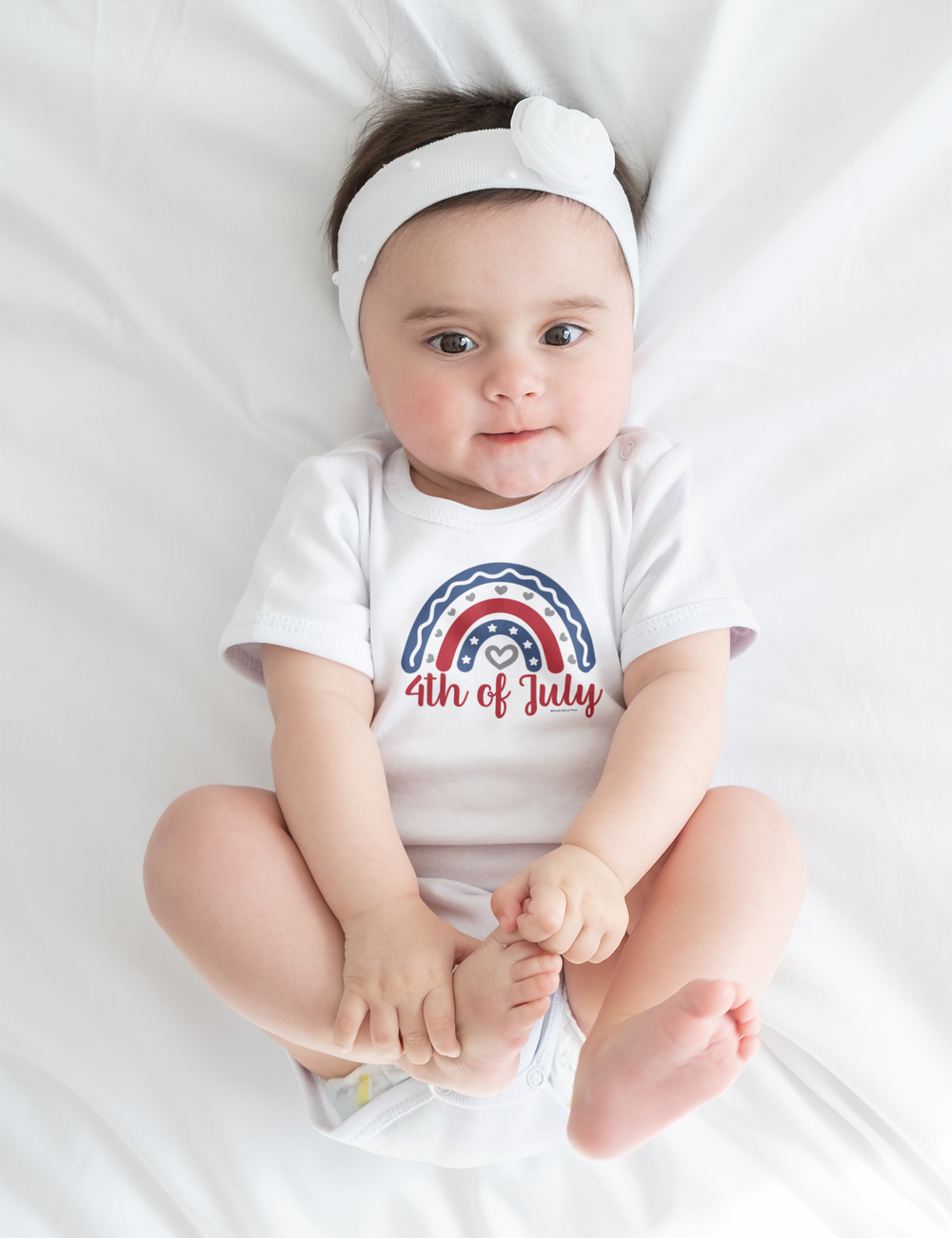A baby lies on a white sheet, wearing a 4th of July Onesie from Worlds Worst Tees. This infant bodysuit is 100% cotton, featuring ribbed knitting for durability and plastic snaps for easy changes.