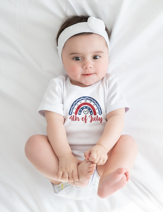 A baby lies on a white sheet, wearing a 4th of July Onesie from Worlds Worst Tees. This infant bodysuit is 100% cotton, featuring ribbed knitting for durability and plastic snaps for easy changes.